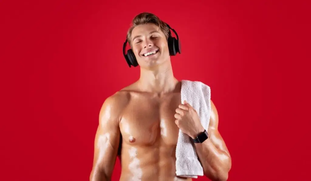 Handsome young sportsman with bare torso and towel listening to music in headphones on red studio background